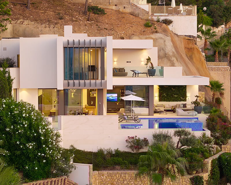 Modern white multi-level villa with a swimming pool, terraces, and large glass windows built into a rocky hillside.