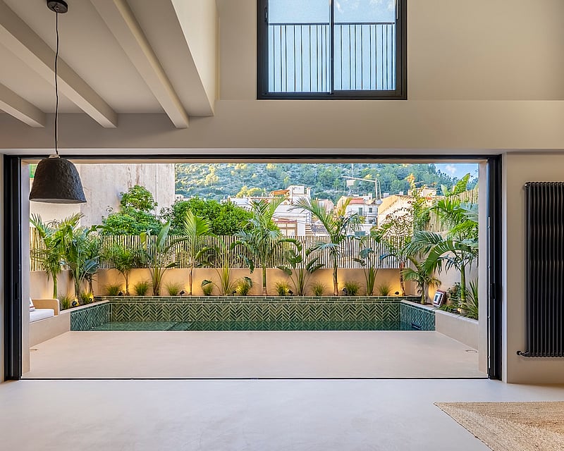 Interior view through a large glass door to a private pool area with tropical plants and green herringbone tiles.