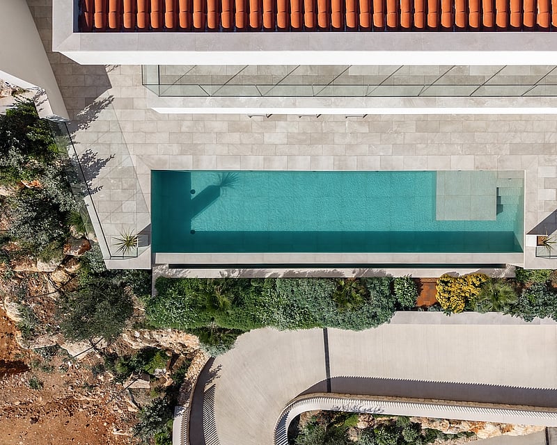 Bird's eye view of a turquoise swimming pool, stone terrace, terracotta roof, and lush Mediterranean landscaping.