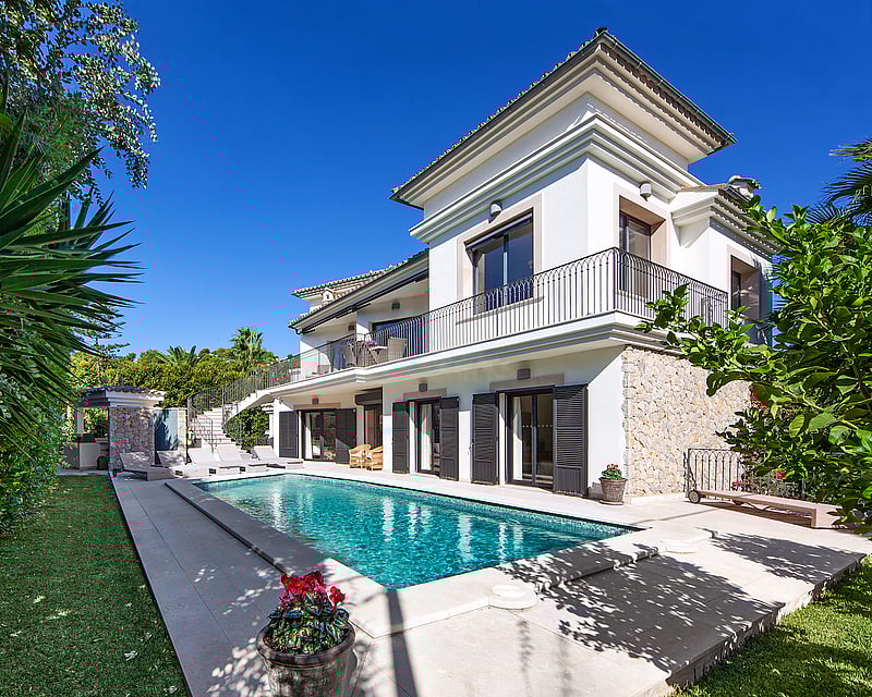 A white two-story villa with a turquoise swimming pool, sun loungers, stone walls, and lush greenery under a clear sky.