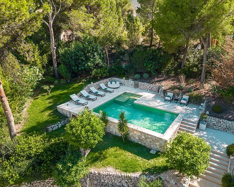 Aerial view of a rectangular turquoise swimming pool with stone deck and white loungers nestled in a pine forest.