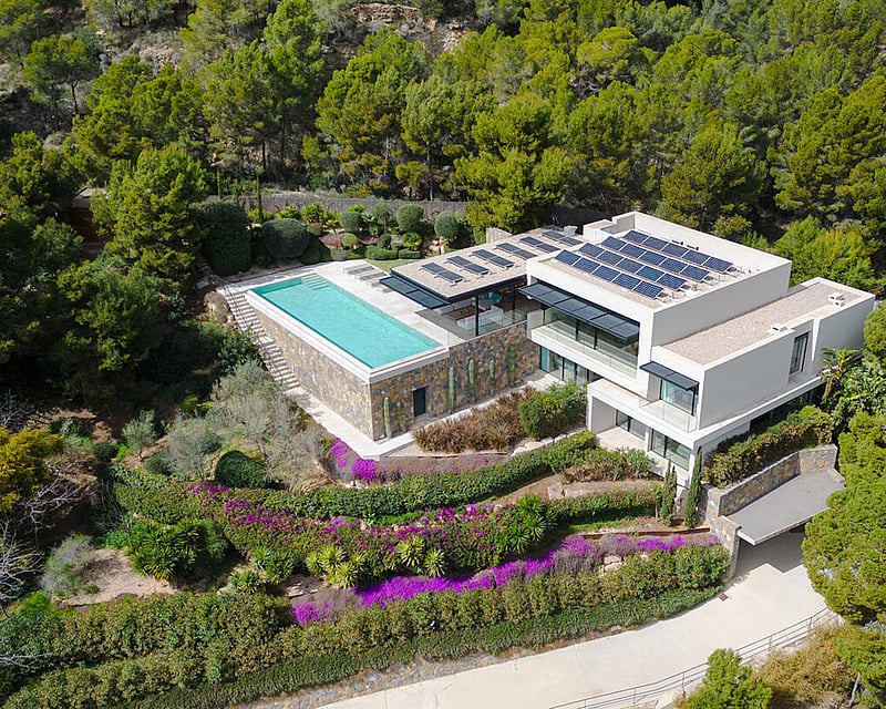Aerial view of a modern white villa with a rectangular infinity pool, solar panels, and surrounding pine forest.