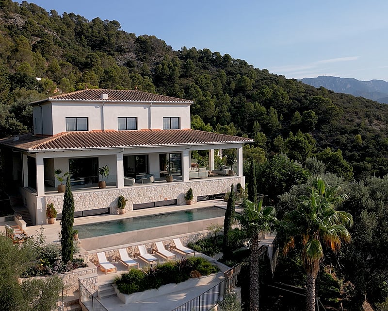 Elevated view of a white two-story villa with a rectangular pool, sun loungers, and lush green mountain backdrop.
