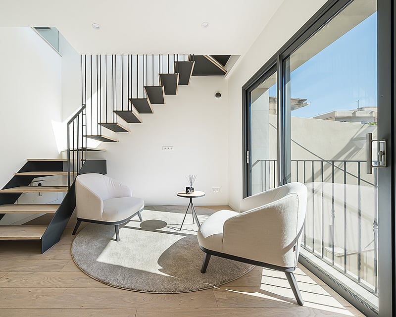 Modern living space featuring a floating wooden staircase, two cream armchairs, and floor-to-ceiling glass doors.