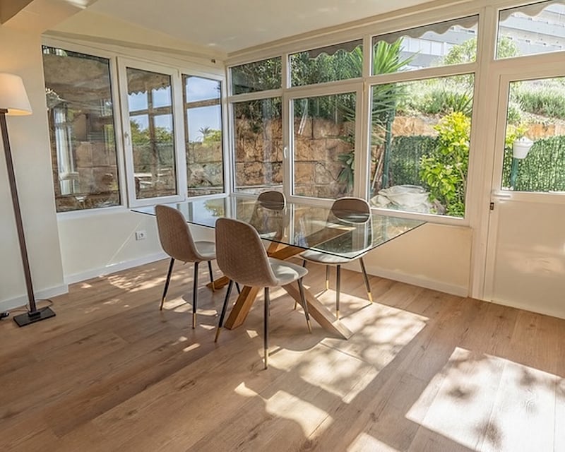 Comedor luminoso con mesa de cristal, sillas tapizadas en beige y amplios ventanales que dan a un jardín mediterráneo.