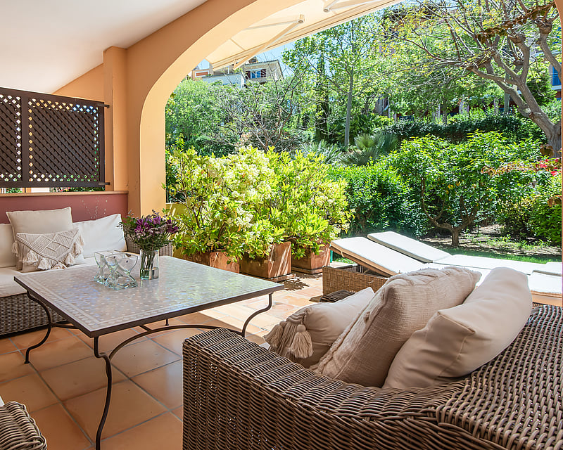 A covered terrace with wicker sofas, cream cushions, and a mosaic table overlooking a vibrant green garden.