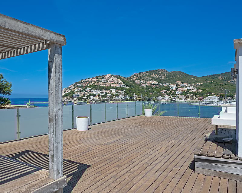 Large wooden rooftop terrace with a pergola overlooking a Mediterranean harbor and hillside villas under a clear blue sky.