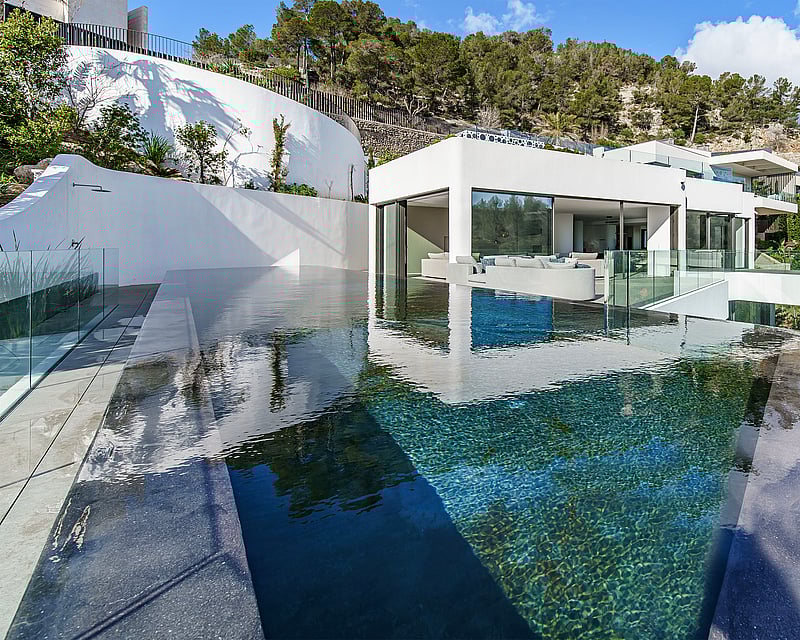 Modern infinity pool with clear blue water overlooking a white minimalist villa nestled in a green hillside.