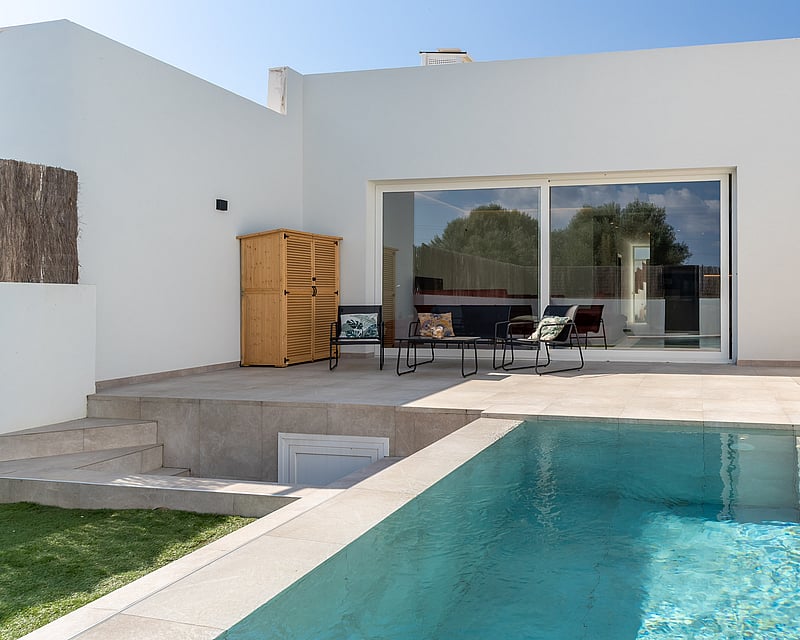 Rectangular turquoise swimming pool with stone steps integrated into a light stone terrace next to a white villa facade.
