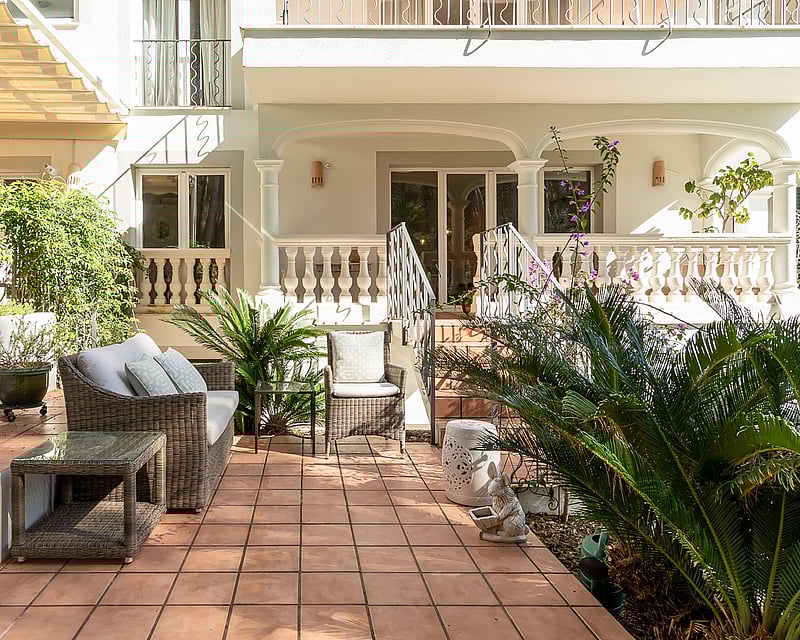 Sun-drenched terracotta tile terrace with wicker lounge furniture and lush greenery against a classic white villa facade.