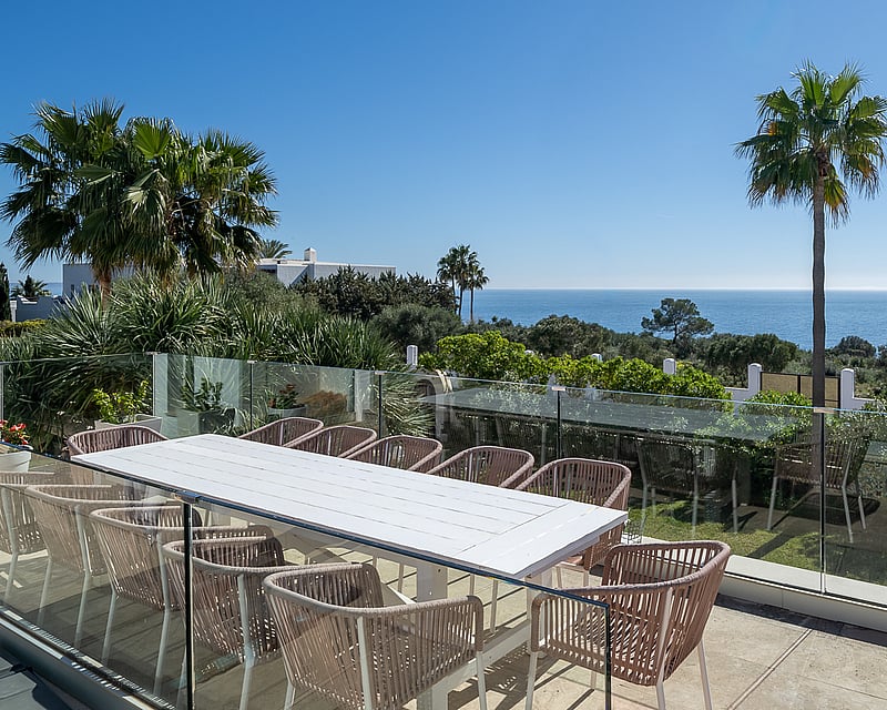 A spacious outdoor terrace with a large white table, stylish chairs, and a glass railing overlooking the sea and palm trees.