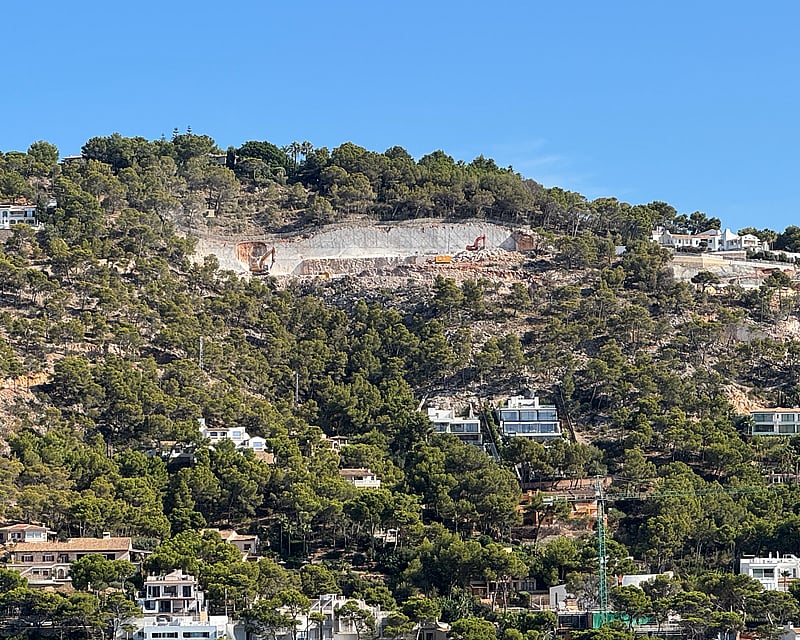 View of a lush green hillside dotted with modern white villas and an active construction site under a clear blue sky.