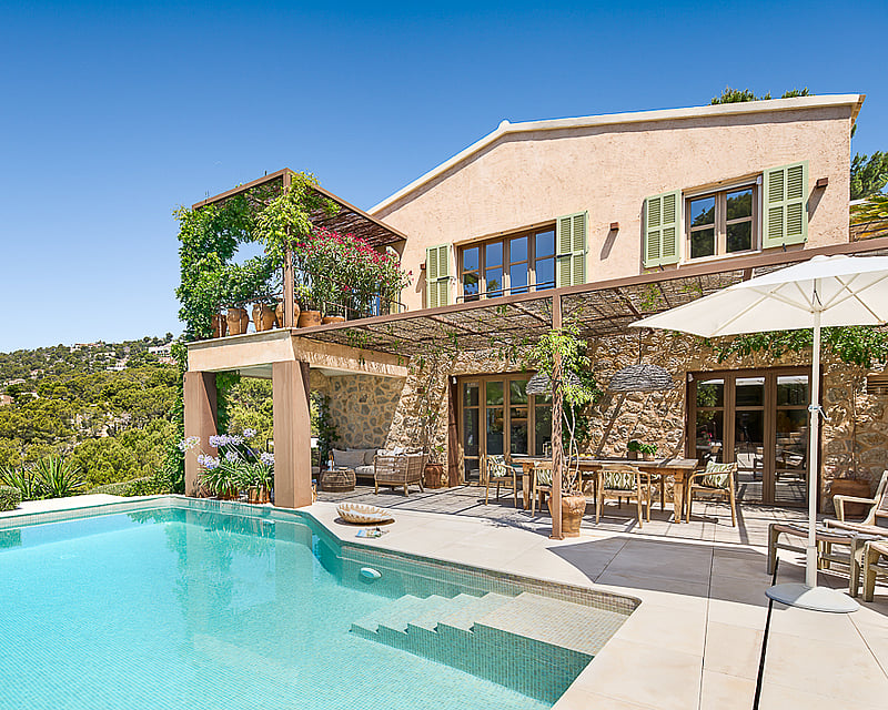 Exterior view of a stone villa featuring a turquoise pool, outdoor dining area, and panoramic hillside views.