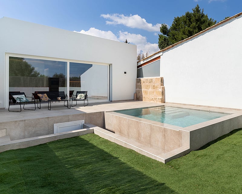 Raised square plunge pool finished in beige stone, adjacent to a white minimalist villa and green lawn.