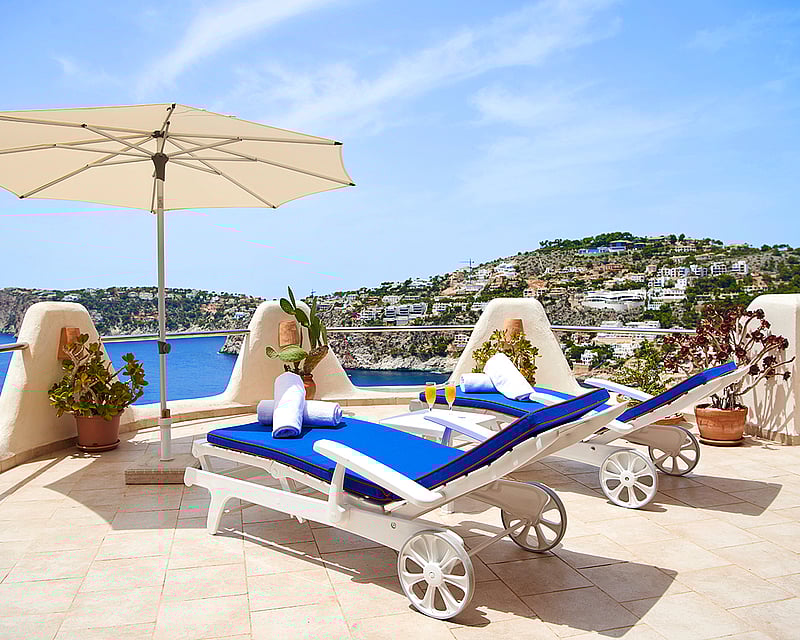 Two blue sun loungers and a white umbrella on a stone terrace overlooking the Mediterranean sea and coastline.