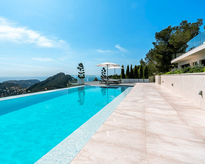 Turquoise infinity pool overlooking the sea and mountains, surrounded by light stone tiles and white umbrellas.