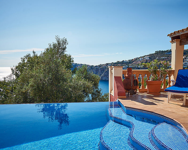 Infinity pool with blue mosaics next to a terracotta terrace overlooking the Mediterranean sea and coastline.