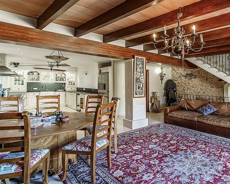 An open-plan living room with dark wooden ceiling beams, a stone wall, leather sofa, and an ornate rug.