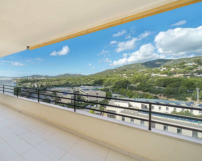 Spacious white tiled balcony with a black metal railing overlooking green hills and a blue sky with white clouds.