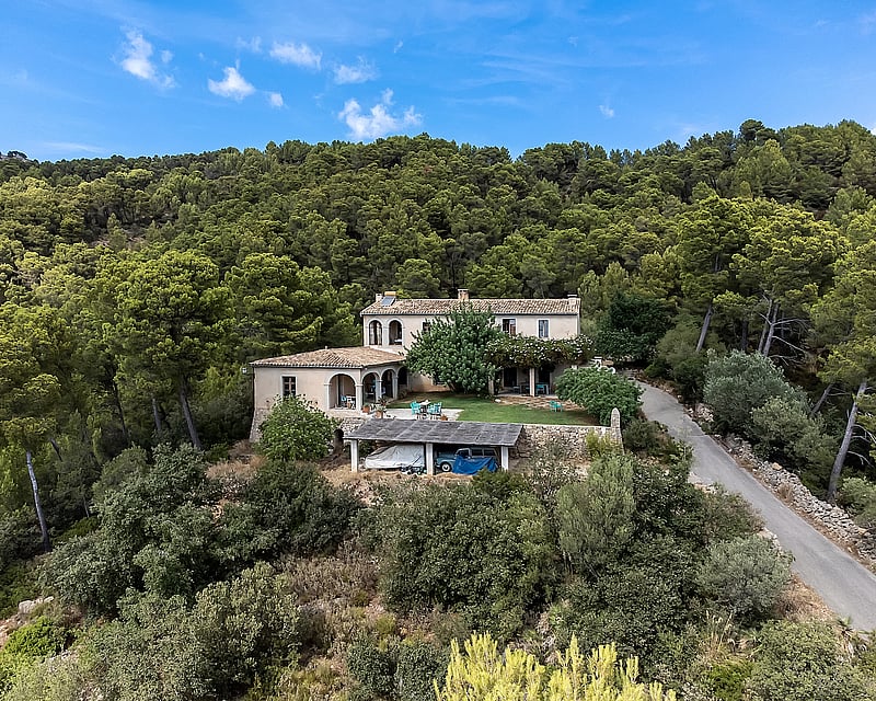 Exterior view of a rustic villa with terracotta roof surrounded by dense pine forest, featuring a lawn and covered parking.
