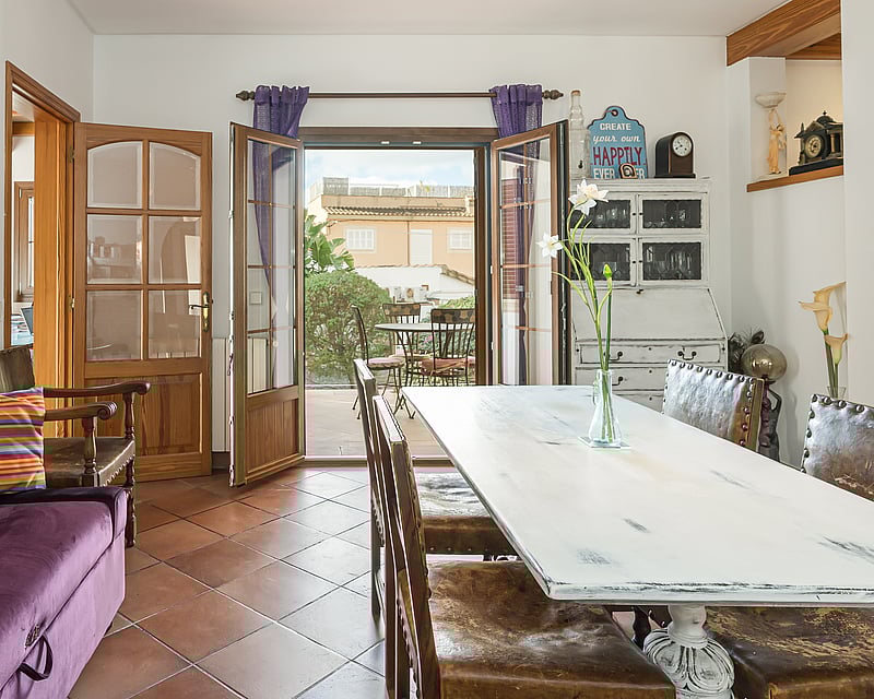 Interior dining room featuring terracotta tile floors, a rustic white wooden table, and open doors leading to a sunny patio.