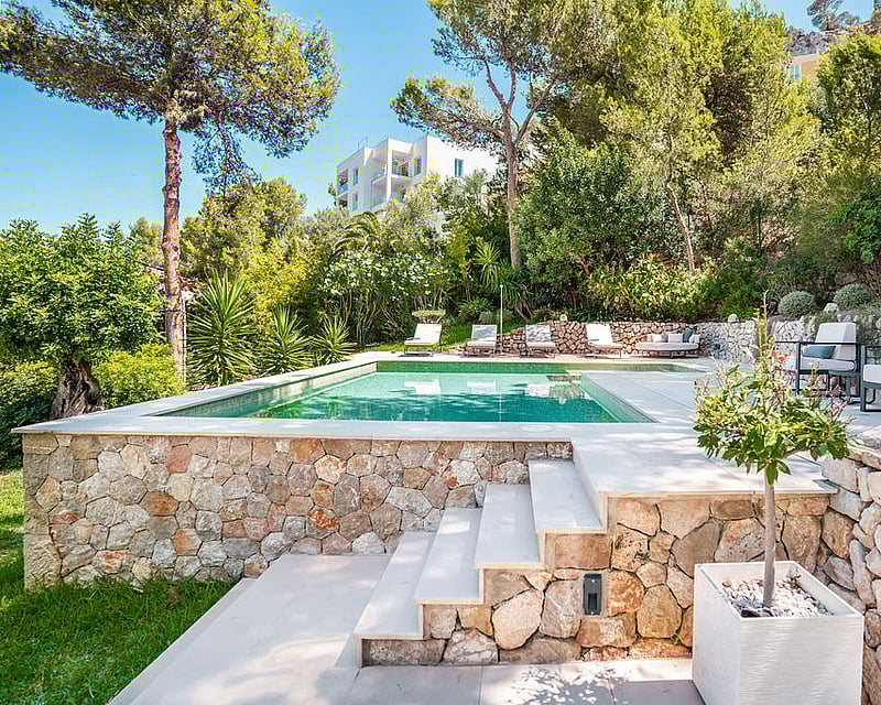 Elevated swimming pool with light stone deck, surrounded by dry stone walls and lush green Mediterranean pines.