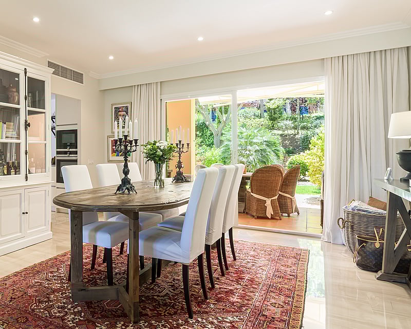Dining room featuring an oval wooden table, white upholstered chairs, and an oriental rug, overlooking a lush garden.