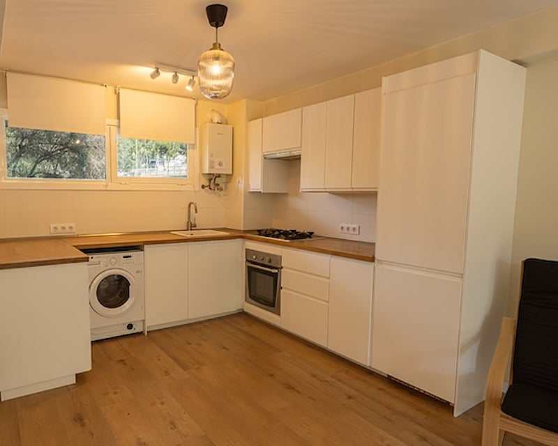 Cocina moderna blanca con encimeras de madera, lavadora integrada, horno y suelos laminados de madera.