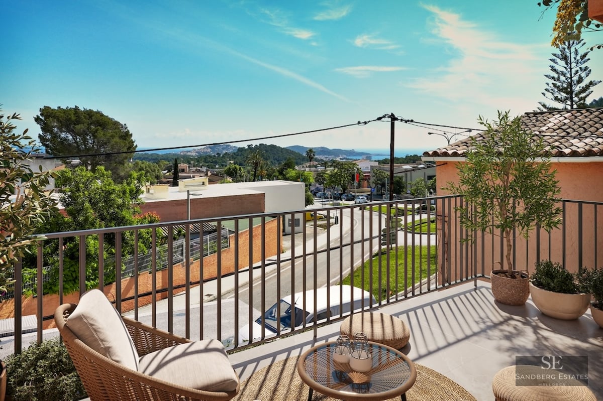 A sunny elevated terrace with a wicker armchair and small table overlooking a coastal town and the sea.