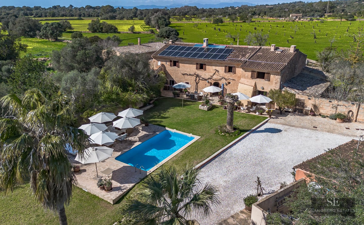 High angle aerial shot of a stone villa with a blue pool, solar panels, and white umbrellas surrounded by green fields.
