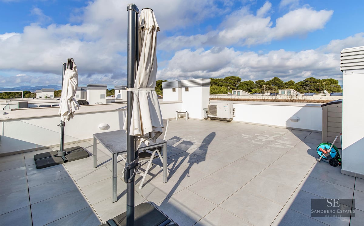Spacious tiled rooftop terrace with grey table, chairs, and two closed white umbrellas under a blue cloudy sky.