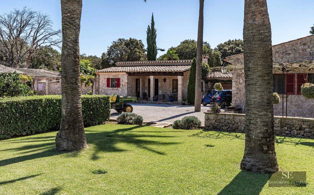 A rustic stone villa with red shutters and a terracotta roof, surrounded by a lush green lawn and palm trees.