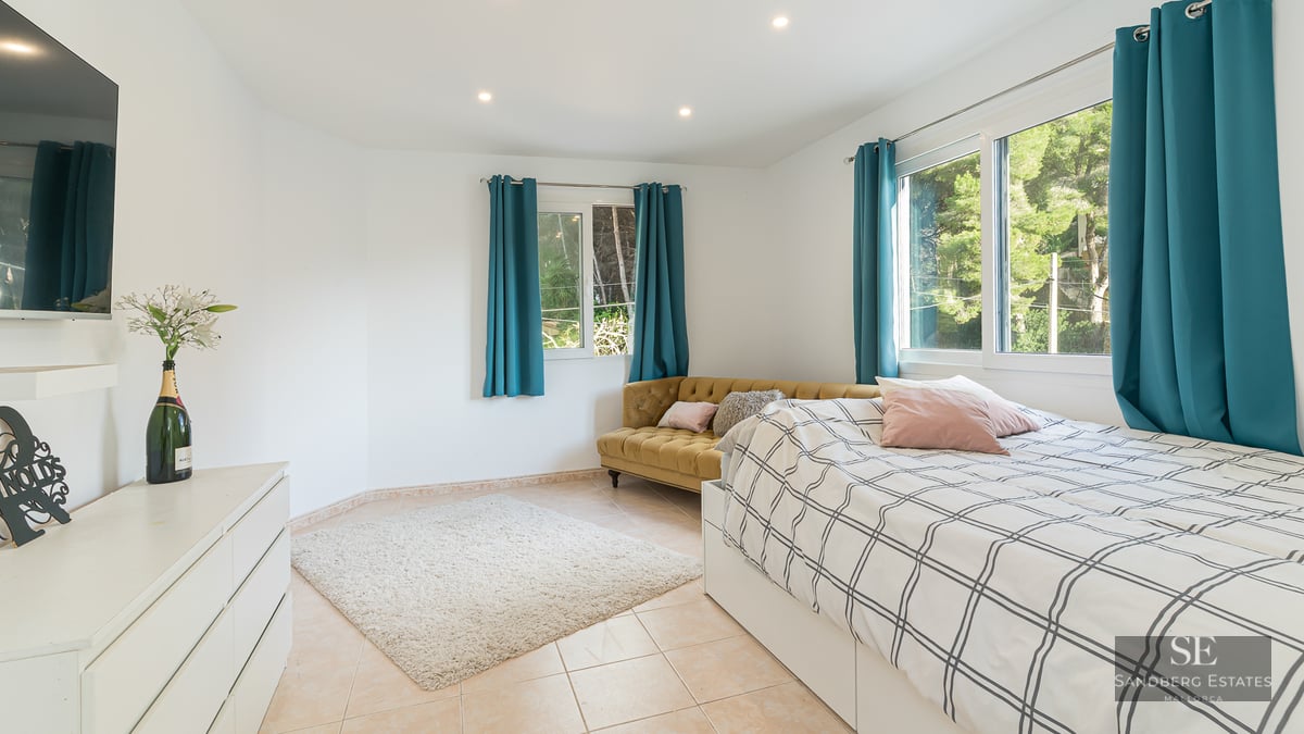 Chambre lumineuse avec lit, canapé en velours moutarde, rideaux bleu canard et vue sur la forêt par les fenêtres.