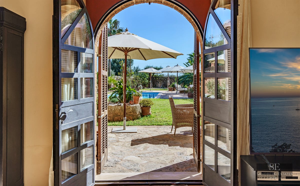 View through open arched wooden doors to a private swimming pool, garden, and white parasols in bright sunlight.