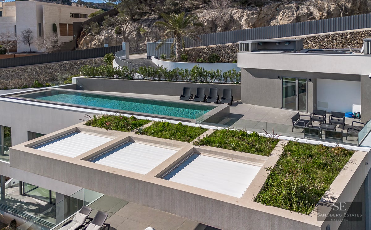 High-angle view of a modern grey villa featuring an infinity pool, sun loungers, and rooftop greenery against a rocky hill.