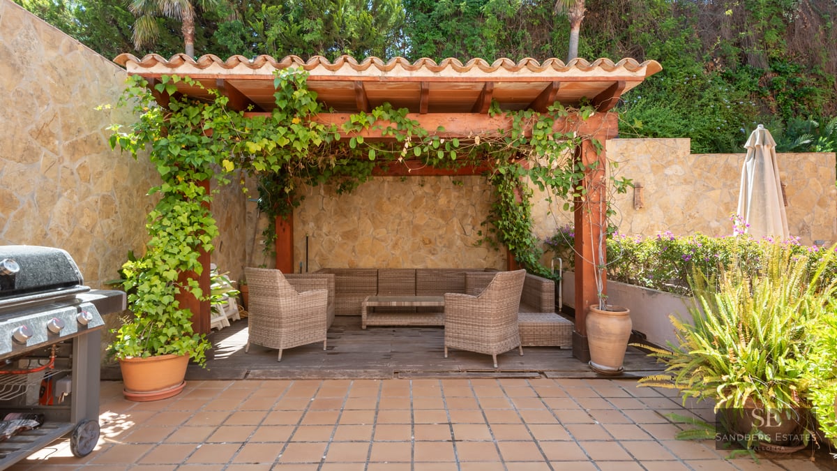 Outdoor terrace featuring a wooden pergola, wicker lounge seating, stone walls, and a grill surrounded by lush plants.