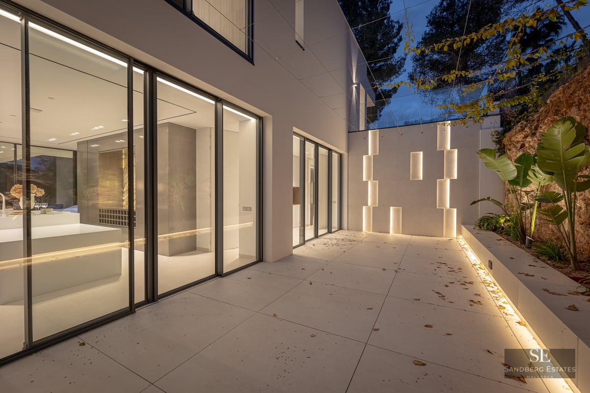 Modern villa terrace at night featuring large glass sliding doors, integrated LED wall lighting, and light stone tiling.