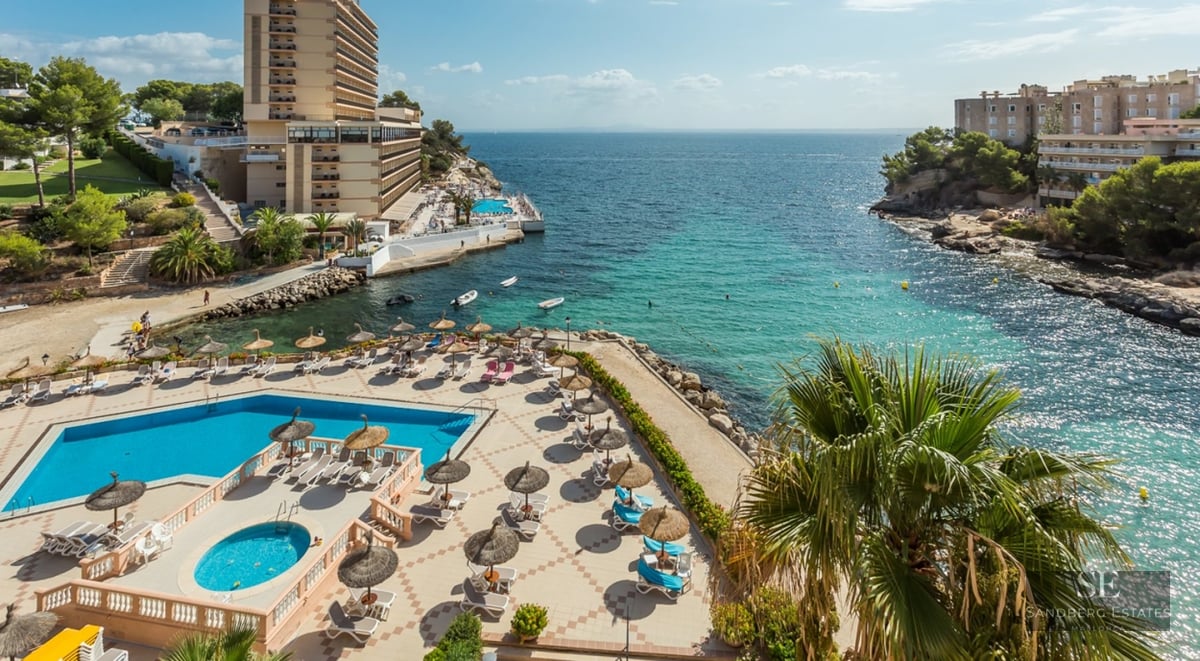 Aerial view of a swimming pool area with sunbeds and umbrellas overlooking a clear turquoise bay and rocky coastline.