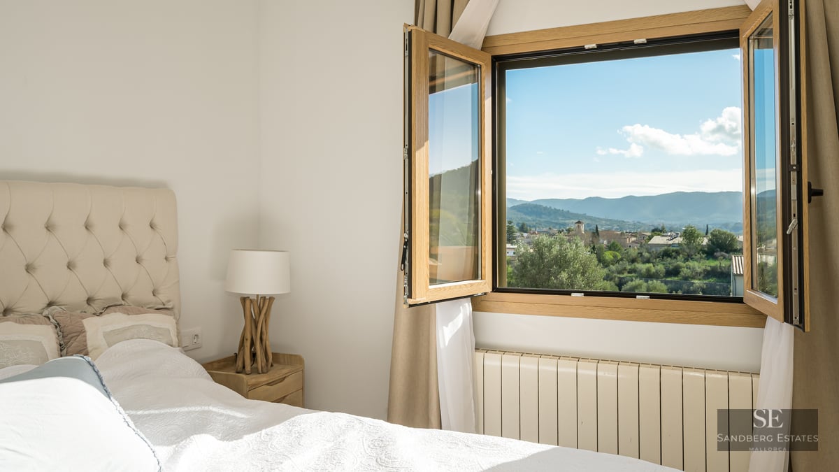 Cozy bedroom with tufted headboard and open window showing panoramic views of a village and mountains.