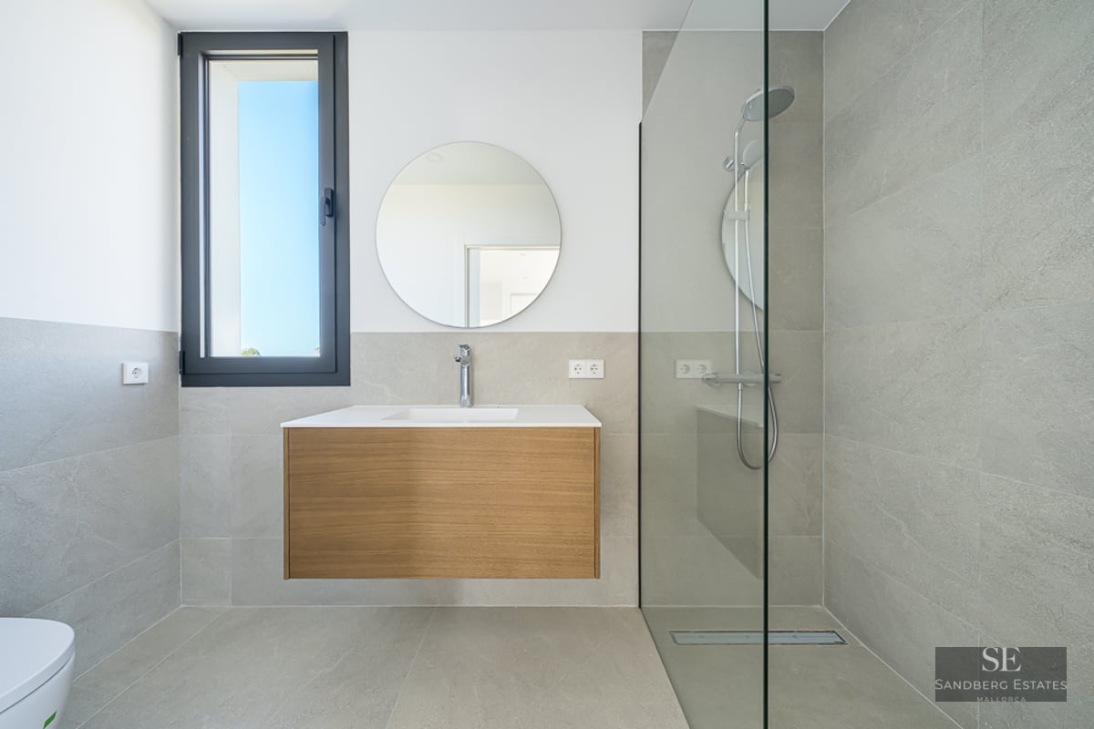 Modern bathroom featuring grey tiling, a floating wooden vanity under a round mirror, and a glass-enclosed walk-in shower.