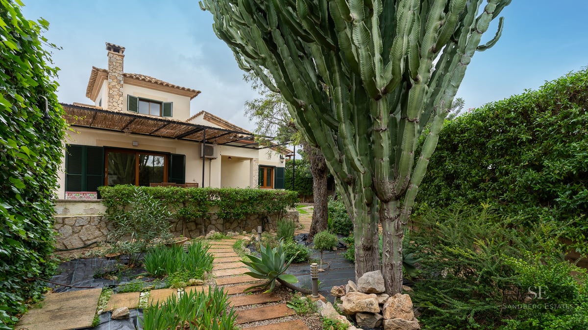View of a lush garden featuring a large cactus, stone pathway, and a Mediterranean home with green shutters.