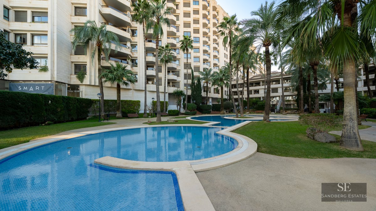 Winding blue swimming pool surrounded by green grass and tall palm trees in front of a modern apartment building.