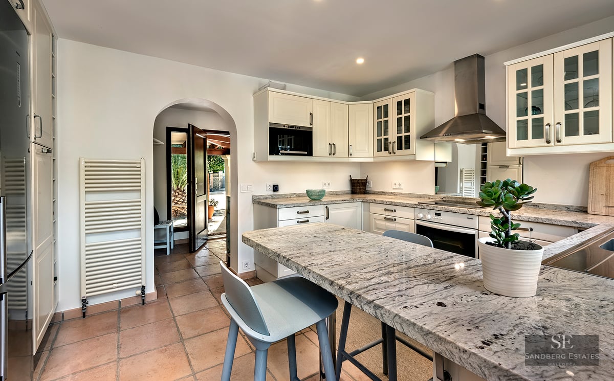 Bright kitchen with white cabinets, granite countertops, terracotta floors, and an archway leading to a sunny exterior.