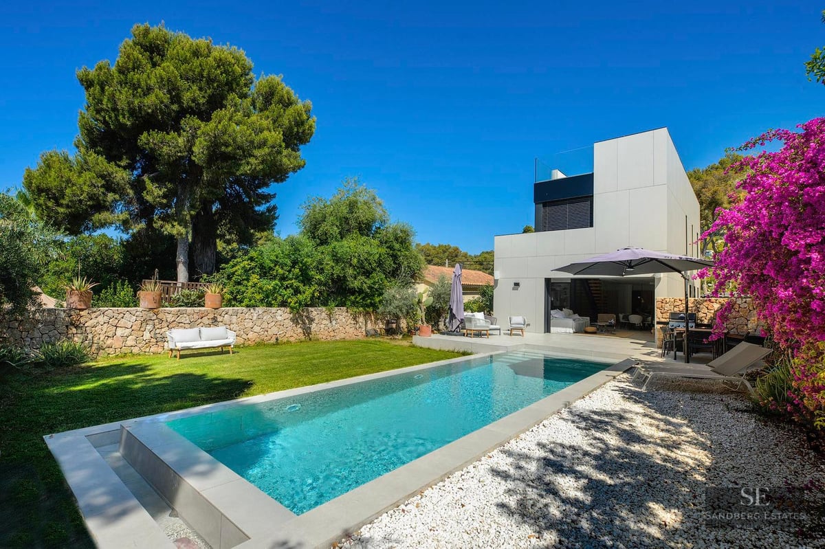 Rectangular swimming pool surrounded by a green lawn, white gravel, and a modern white villa under a clear blue sky.