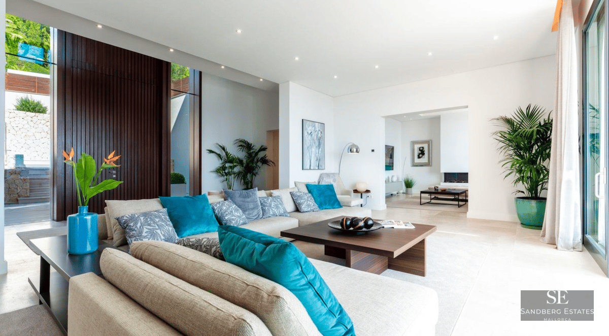 Spacious modern living room with beige sectional sofa, teal pillows, wooden table and floor-to-ceiling windows.