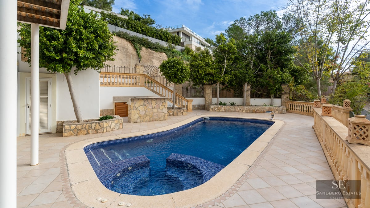 Blue swimming pool with integrated seating on a spacious tiled terrace surrounded by balustrades and trees.