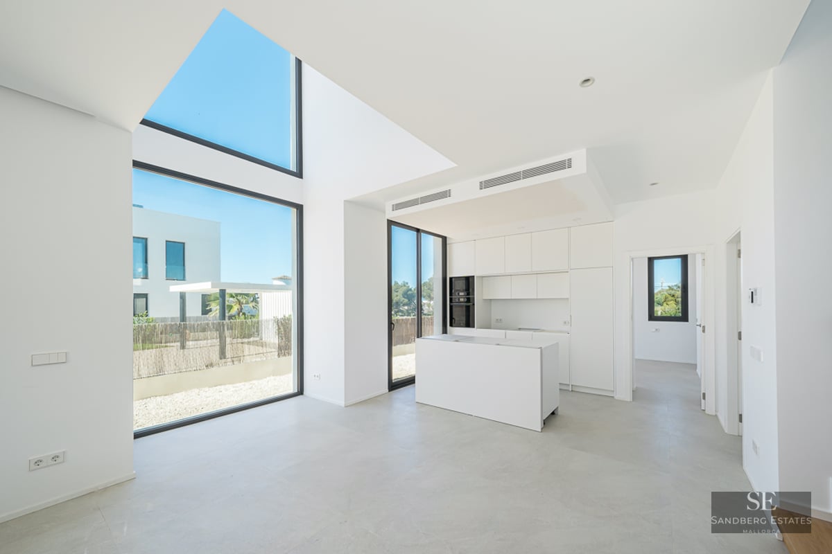 Minimalist white kitchen with an island, double-height floor-to-ceiling windows, and bright natural light.