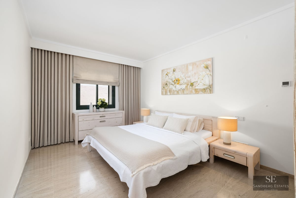 Chambre lumineuse avec lit blanc, tables de chevet en bois, tableau floral et sol en carrelage beige.