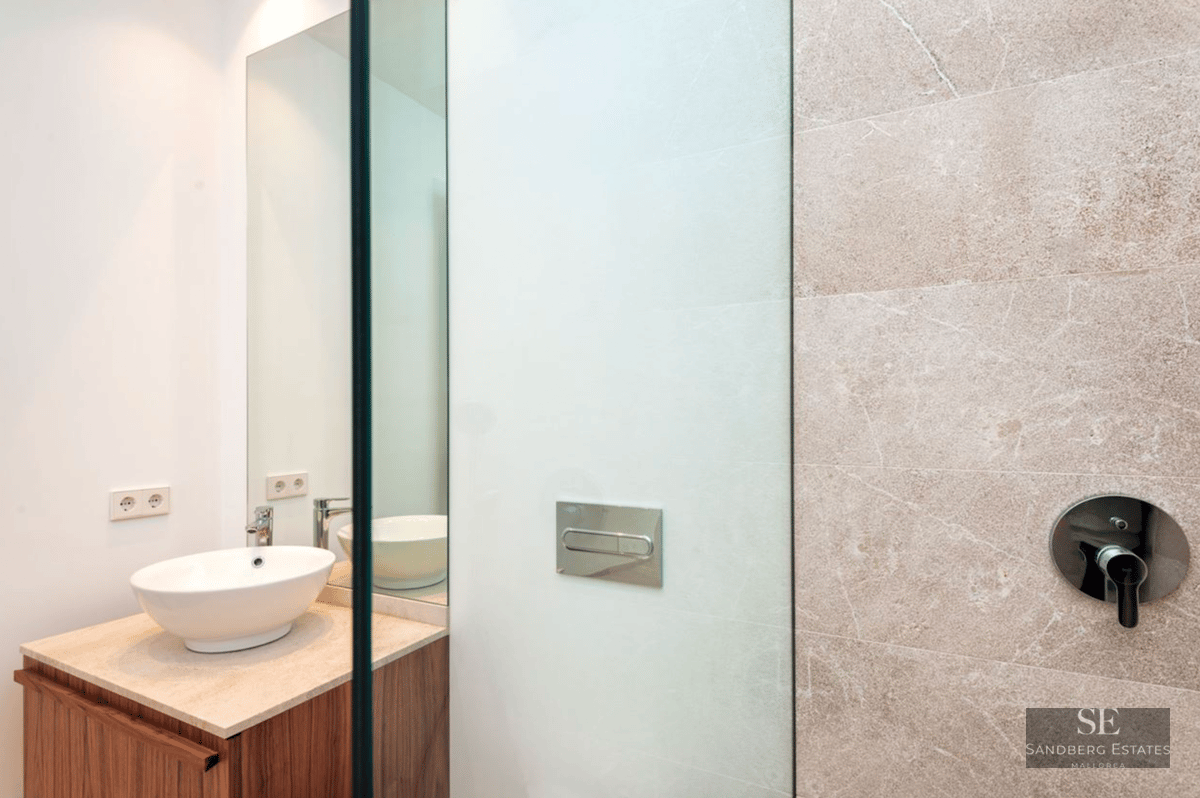 A white vessel sink on a stone counter next to a textured stone wall with a shower mixer and a large mirror.