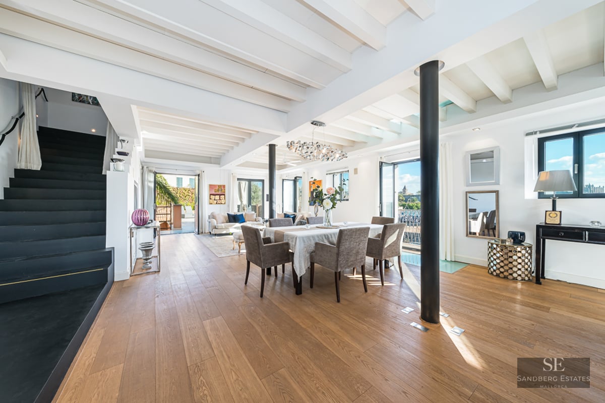 Bright open-plan room with wooden floors, white beamed ceiling, dining set, and a black staircase on the left.